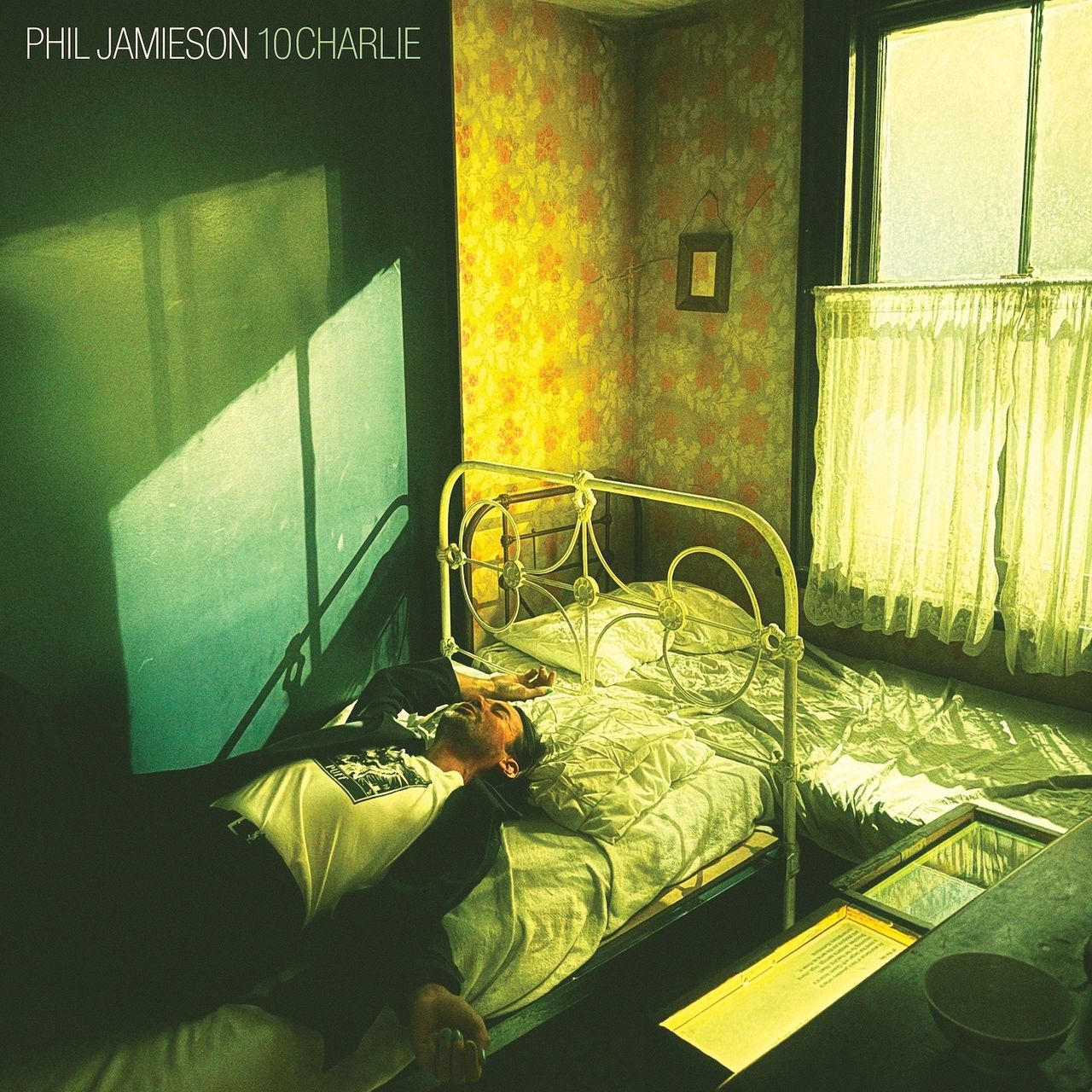 Album cover for Phil Jamieson's 10CHARLIE: a man lies across a metal frame bed in a sunlit room with floral wallpaper and green cast shadows.