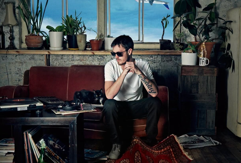 Promotional portrait of Australian rapper 360 sitting on a couch in a room with plants and a cat.