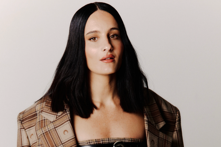 French techno DJ and producer Anetha posing in a plaid jacket with dark straight hair against a neutral background for a press portrait photo.