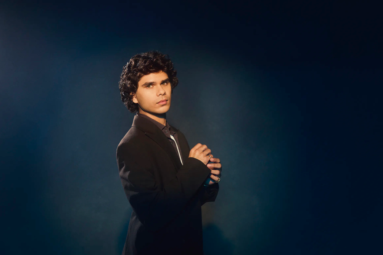 Promotional portrait of Australian R&B singer Budjerah wearing a black suit jacket, posing against a dark blue studio background.