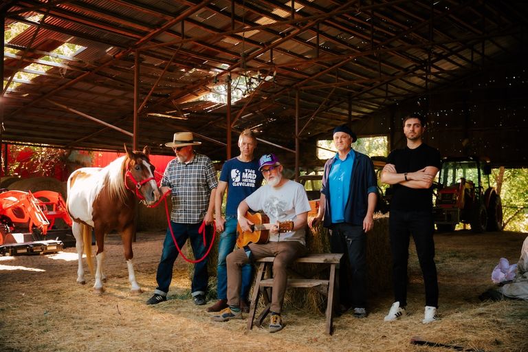 Alt-rock band CAKE standing in a rustic barn with a horse, guitar, and farm equipment.