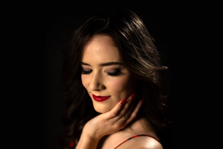 Dramatic portrait of artist Carmen looking down with closed eyes, wearing red lipstick and holding her hand to her cheek.