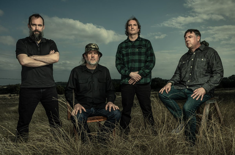 Press photo of American rock band Clutch posing outdoors in a grassy field under a cloudy sky.