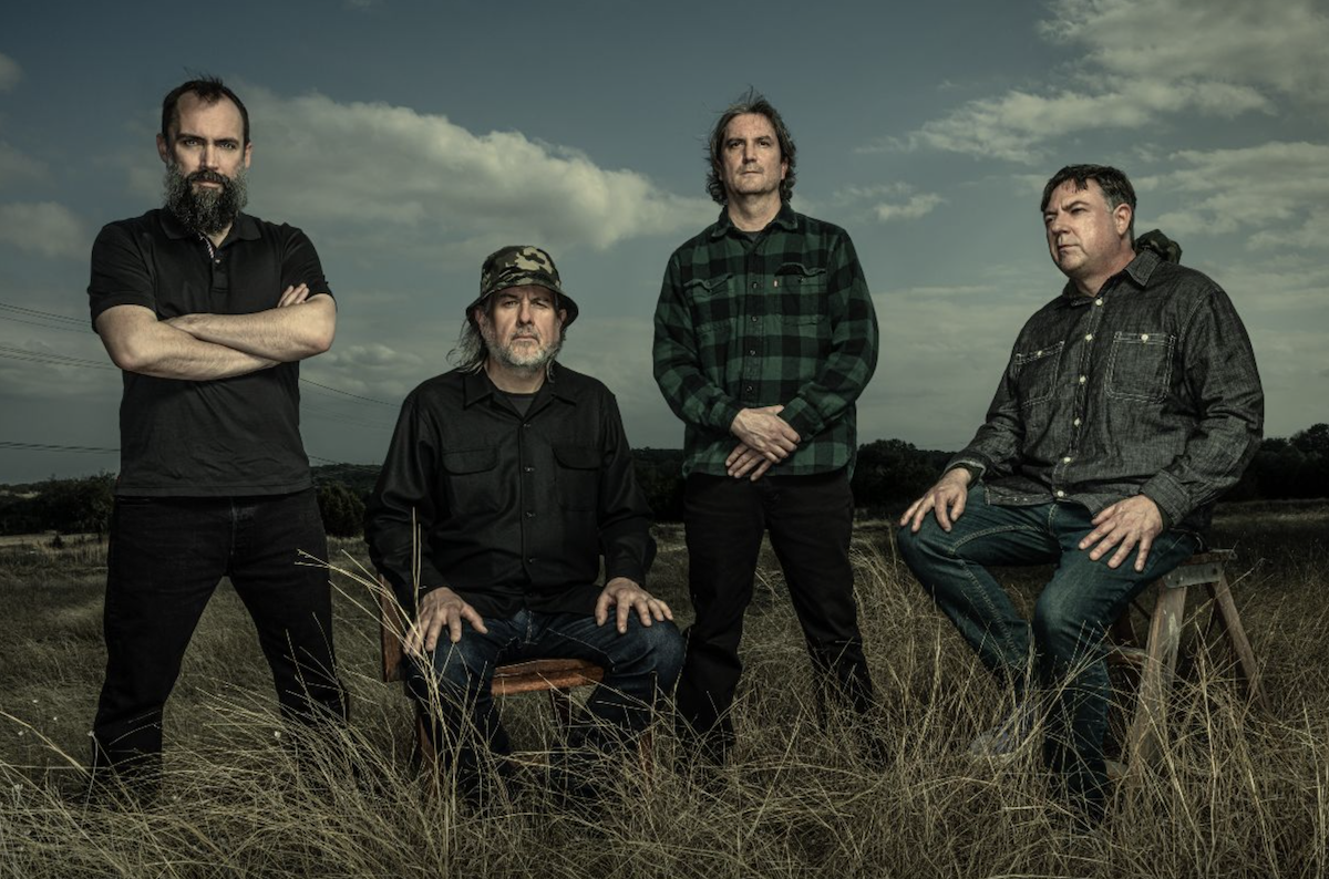 Press photo of American rock band Clutch posing outdoors in a grassy field under a cloudy sky.