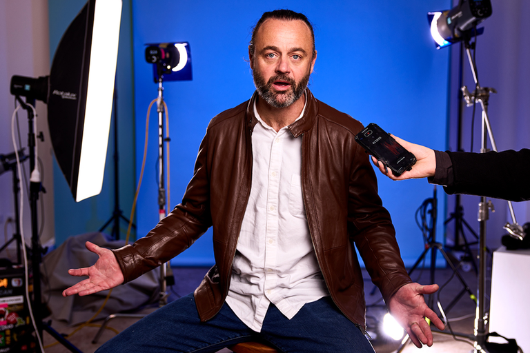 Actor in the play Commentary being interviewed with a smartphone in a studio setting with lighting equipment and a blue background.