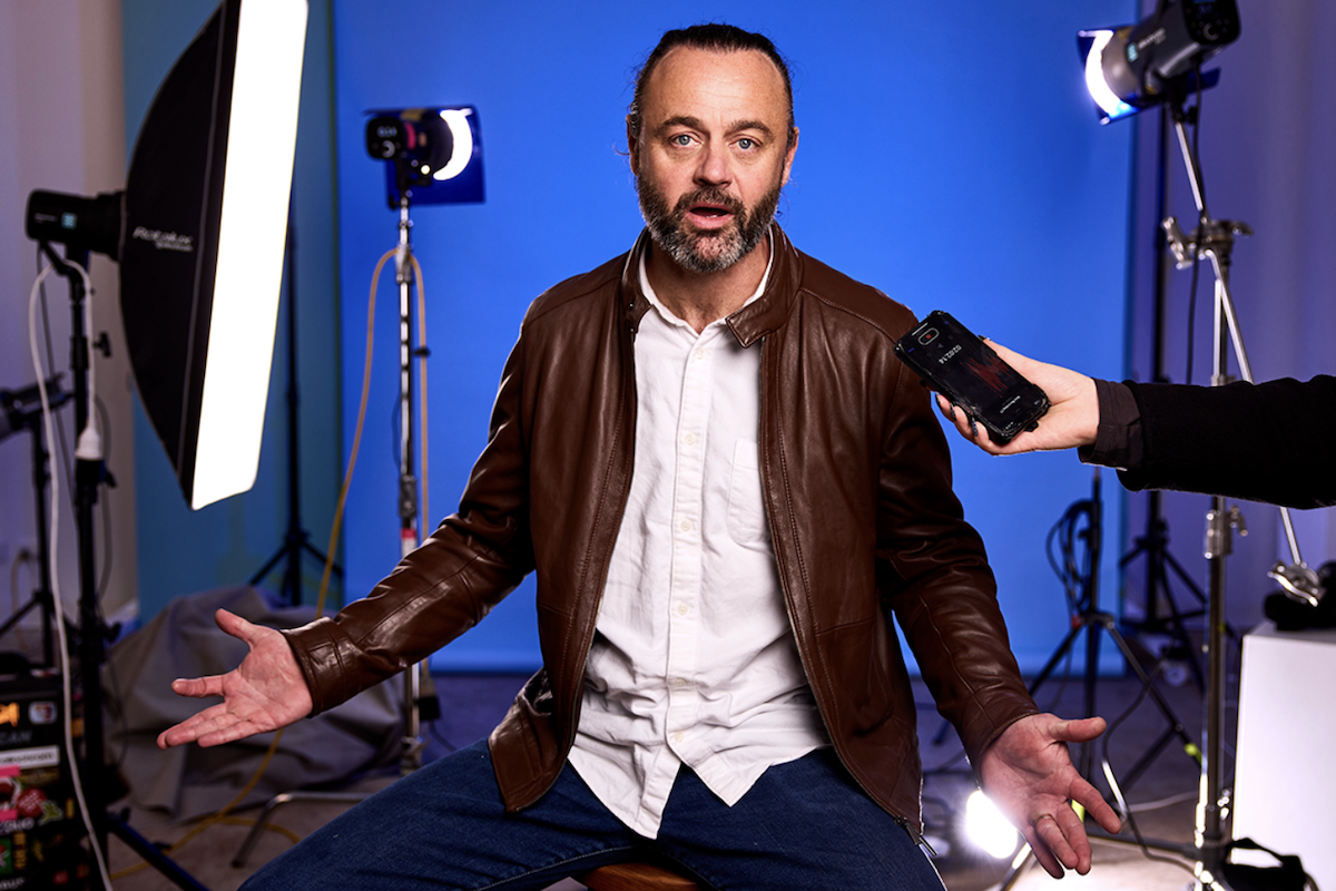 Actor in the play Commentary being interviewed with a smartphone in a studio setting with lighting equipment and a blue background.