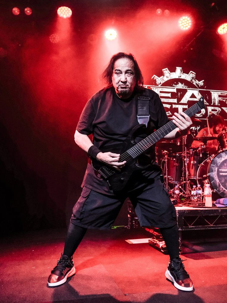 Fear Factory guitarist Dino Cazares playing a black 7-string electric guitar live on stage under red lighting with the band logo background.