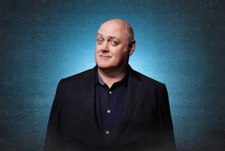 Irish comedian and television presenter Dara Ó Briain smiling in a black suit jacket and blue button-up shirt against a starry blue background.