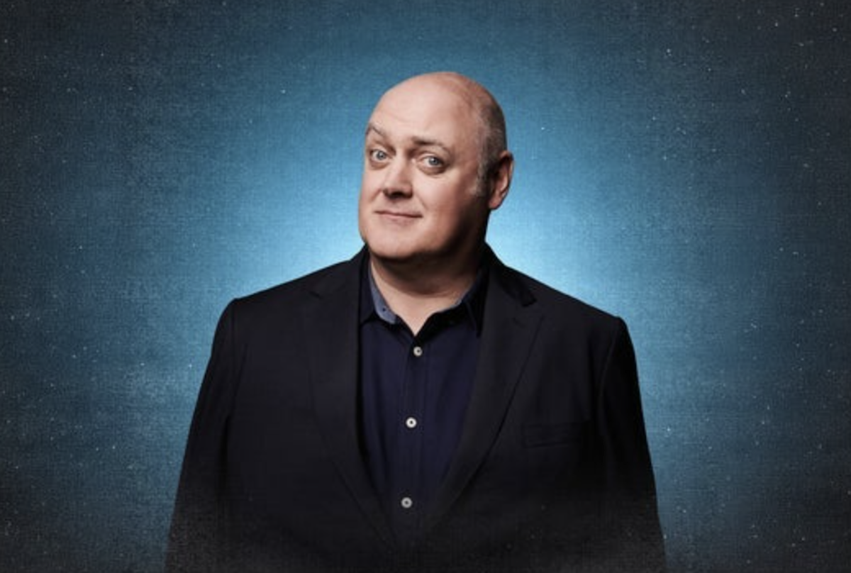 Irish comedian and television presenter Dara Ó Briain smiling in a black suit jacket and blue button-up shirt against a starry blue background.