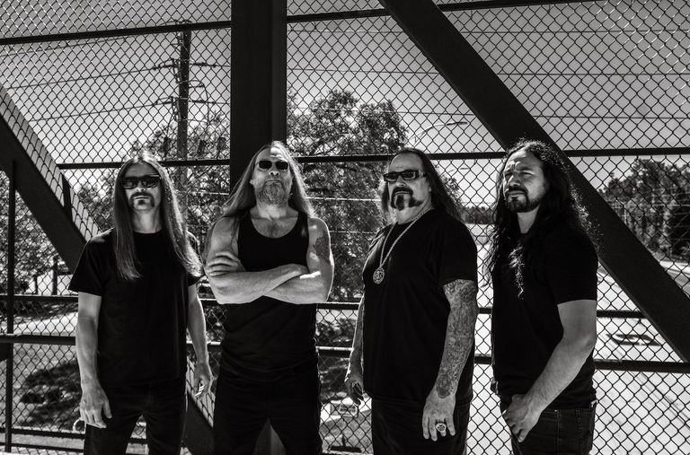 Black and white portrait of American death metal band Deicide members posing together outdoors in front of a chain-link fence structure