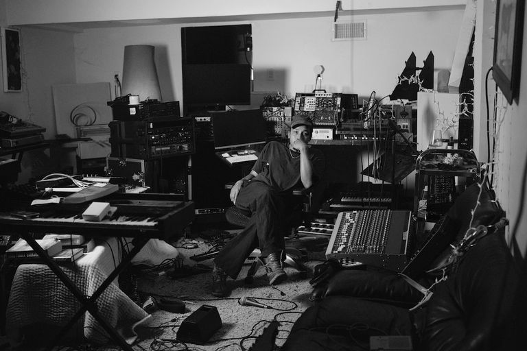 Black and white portrait of musician Dijon sitting in a home recording studio surrounded by keyboards, mixing boards, and audio equipment.