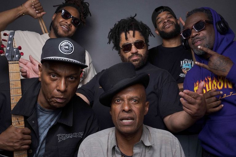 Group photo of ska-punk band Fishbone posing together, featuring a man in a black top hat in the center front and another holding a guitar.
