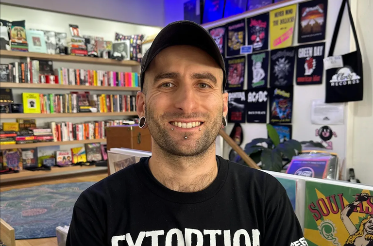 Clarity Records owner Footy smiling inside his Adelaide record store surrounded by band posters, books, and vinyl records for a promo portrait.