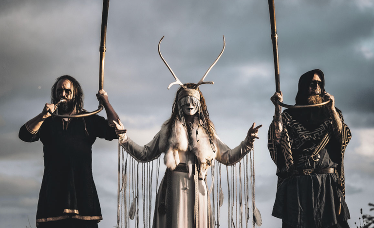 Experimental folk music group Heilung performing outdoors in traditional pagan attire and antlers.