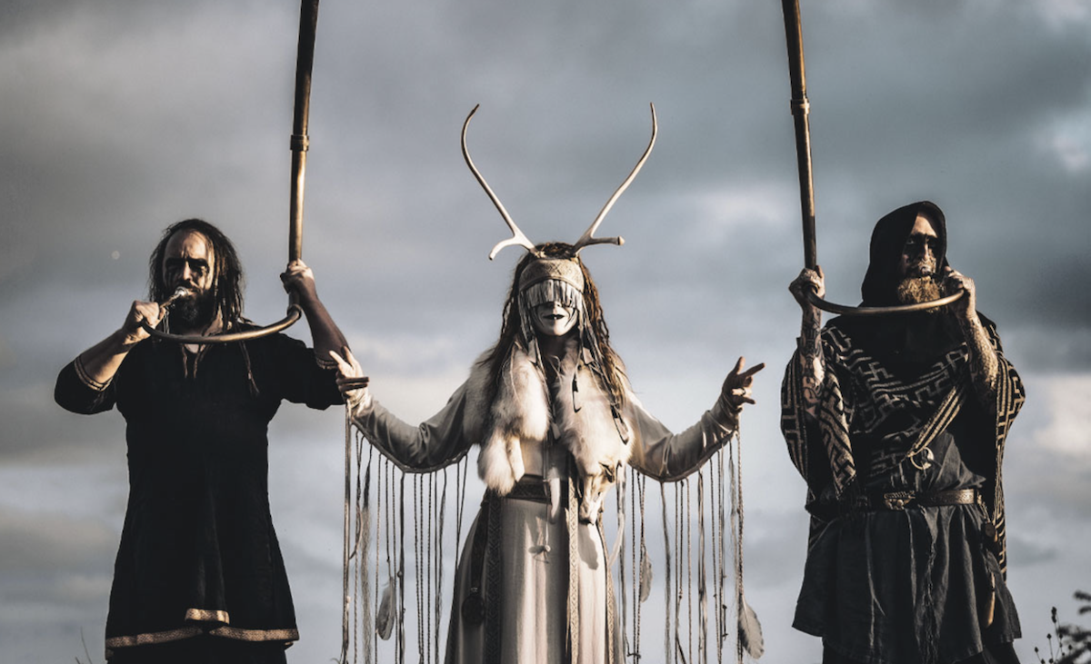 Experimental folk music group Heilung performing outdoors in traditional pagan attire and antlers.