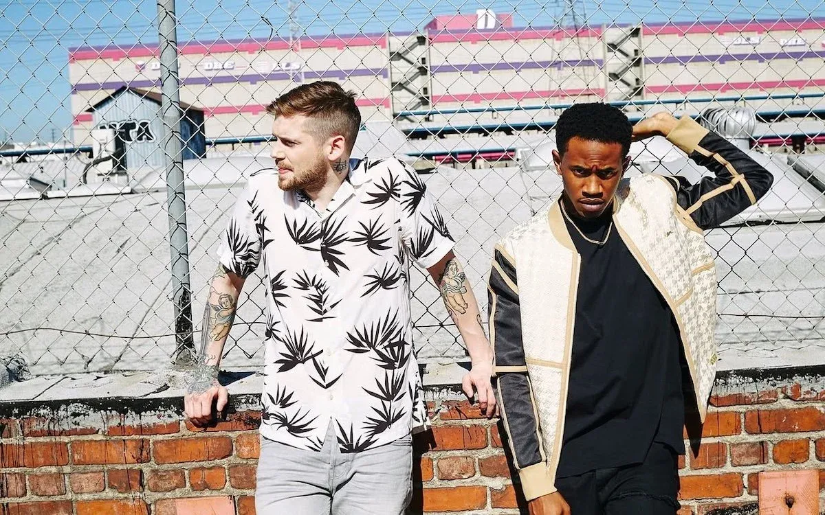 American pop duo MKTO, Malcolm Kelley and Tony Oller, posing in front of a chain-link fence on an urban rooftop portrait.