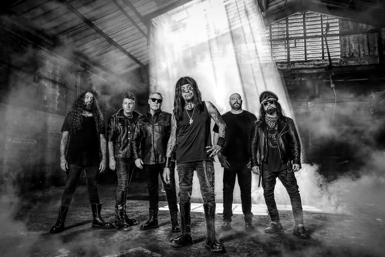 Industrial metal band Ministry standing in a smoky, dark warehouse location with light beams shining down in a black-and-white portrait photo.