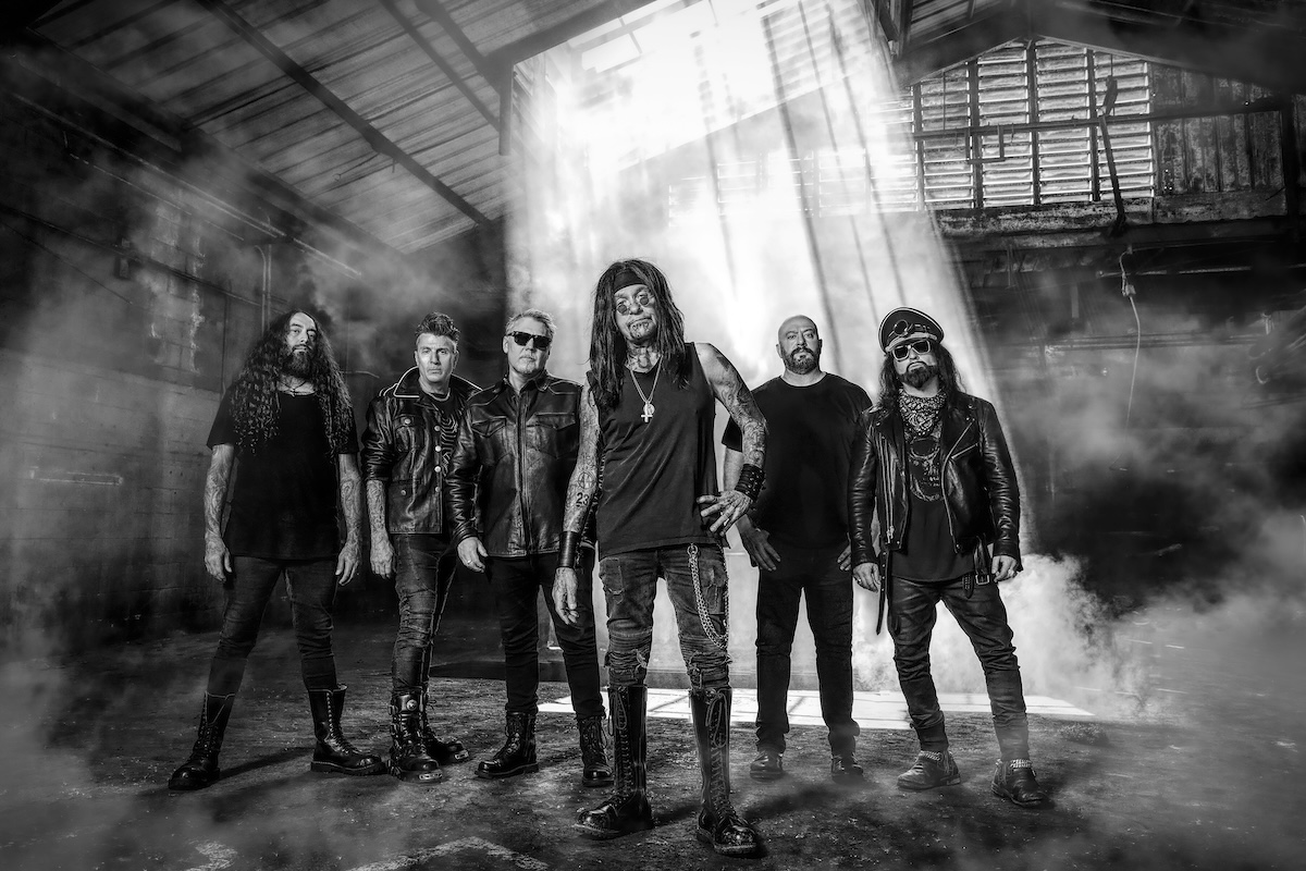 Industrial metal band Ministry standing in a smoky, dark warehouse location with light beams shining down in a black-and-white portrait photo.