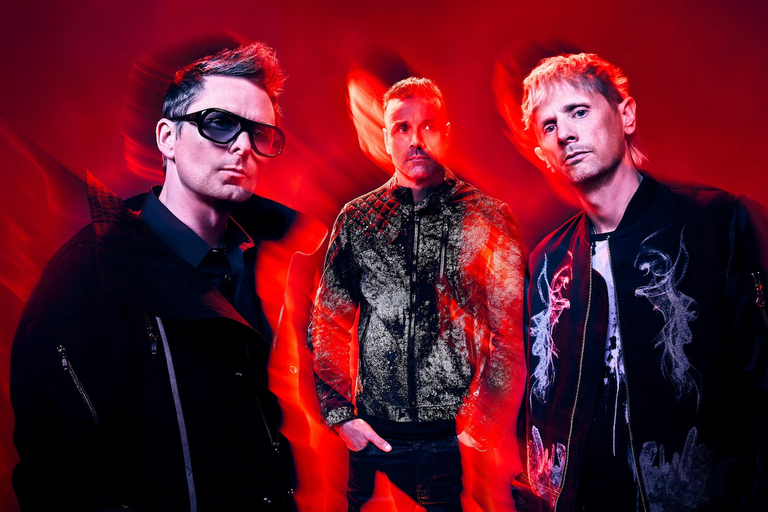 English rock band Muse posing in dark jackets against a vibrant red backdrop with dramatic light streaks.