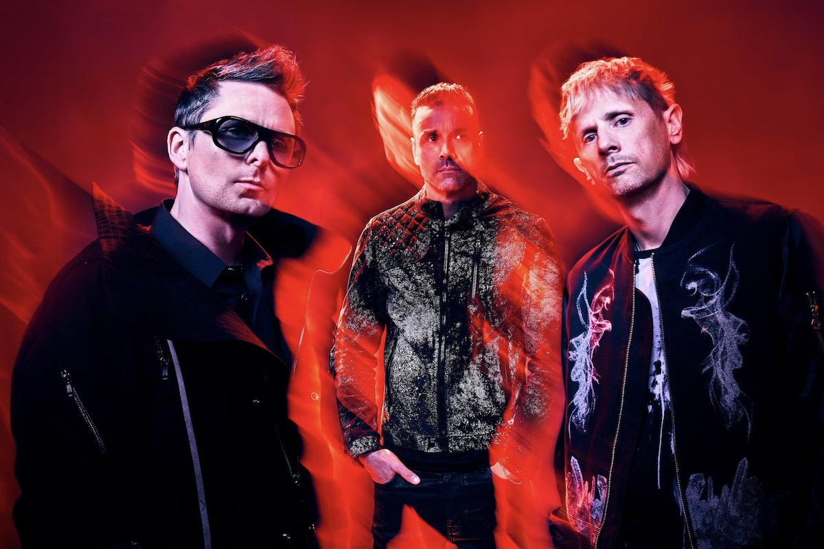 English rock band Muse posing in dark jackets against a vibrant red backdrop with dramatic light streaks.
