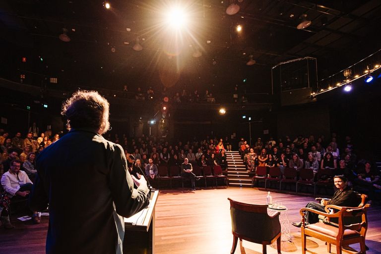 Speaker from behind at a podium addressing an audience in a dim, spotlighted theater room at a Music SA industry event
