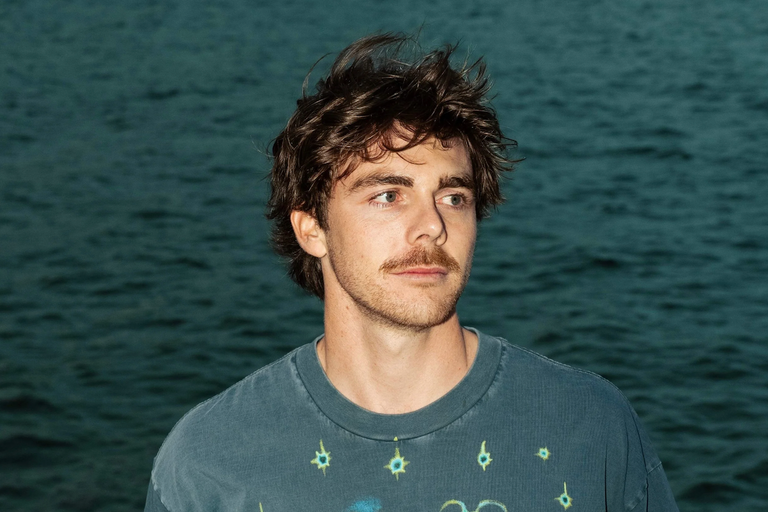 Promotional headshot portrait of musician Newport with a mustache, looking off-camera outdoors against a dark ocean water background.
