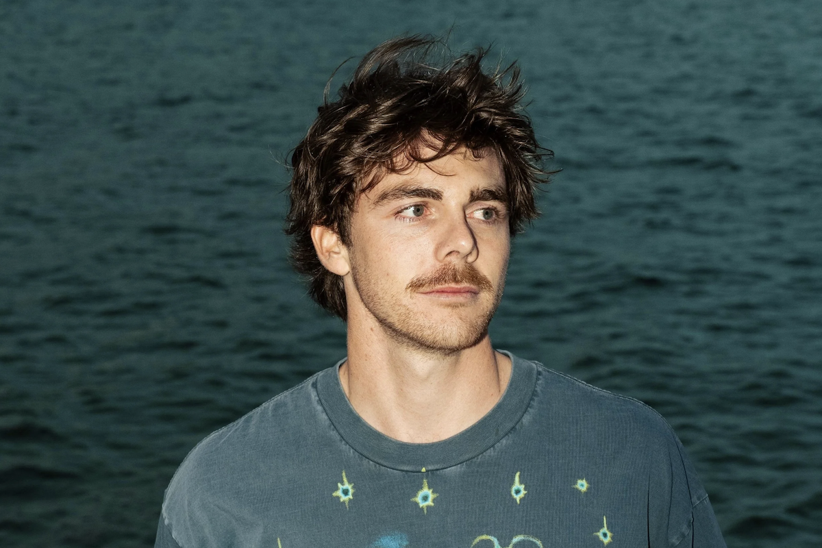Promotional headshot portrait of musician Newport with a mustache, looking off-camera outdoors against a dark ocean water background.