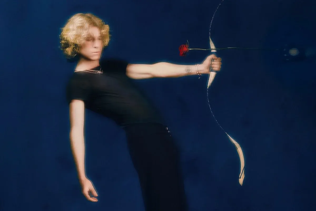 Musician Noah Hill posing with a bow and aiming a red rose like an arrow against a deep blue background in a press photo.