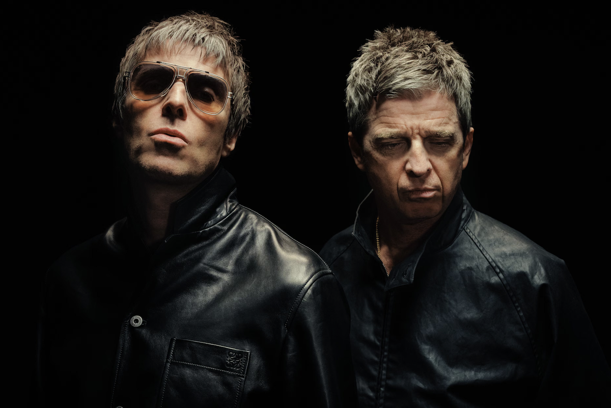 English rock band Oasis brothers Liam and Noel Gallagher wearing black jackets in a moody studio portrait photo.