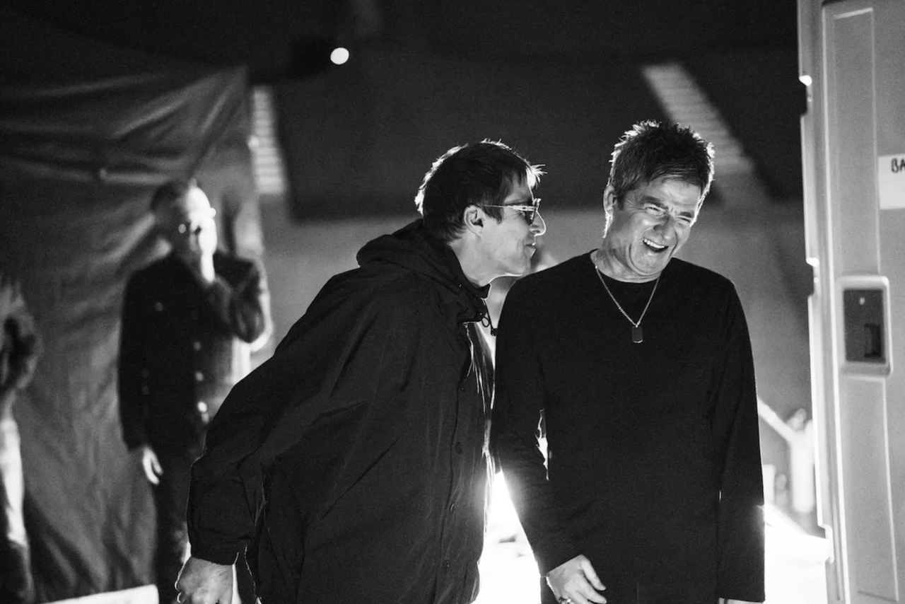 British rock band Oasis members Liam and Noel Gallagher laughing backstage in a black-and-white candid press photo.