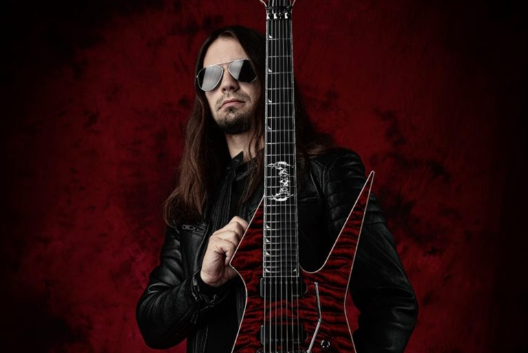 Steffen Kummerer of technical death metal band Obscura holding a red flame-top electric guitar, wearing sunglasses against a dark red backdrop.