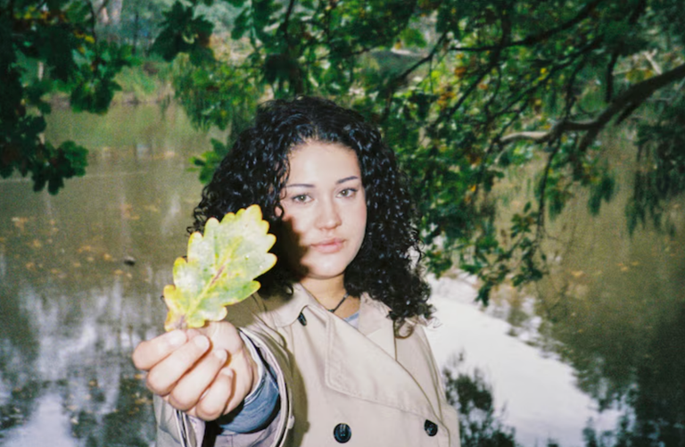 Promotional headshot of Australian musician Odette holding out a yellowing leaf outdoors, wearing a trench coat in front of a river and trees.