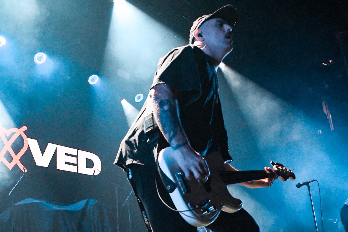 Outloved guitarist performing live on stage in a cap, lit by dramatic blue stage lights with the band logo visible in the background.