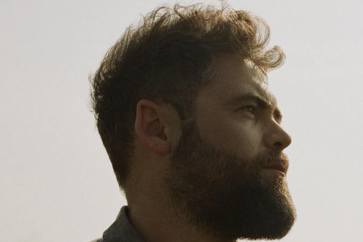 Side profile portrait of singer-songwriter Passenger (Mike Rosenberg) with a beard and curly hair, looking upward against a soft outdoor sky.
