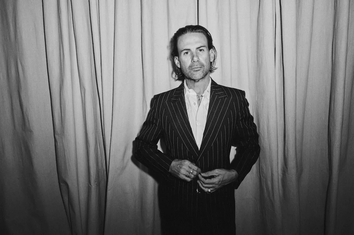 Black and white portrait of Australian musician Phil Jamieson wearing a pinstripe suit, adjusting his jacket in front of a curtain background.