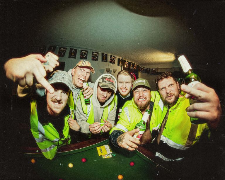 Heavy music group PINTGLASS wearing high-vis vests and holding beer bottles around a pool table.