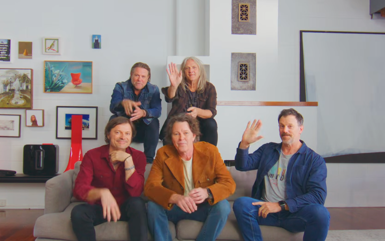 Australian rock band Powderfinger members posing together indoors on a couch and smiling at the camera, some waving hello
