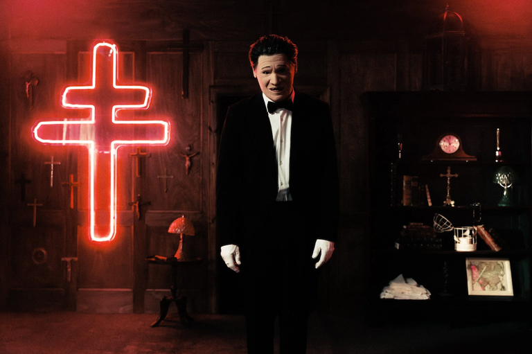 Rock group President frontman in a mask and tuxedo standing in front of a glowing red cross in a promotional photo.