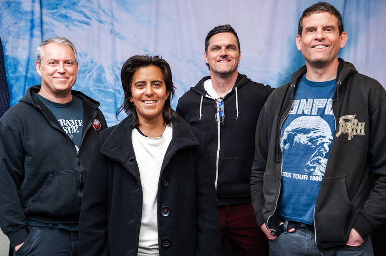 The four members of the punk rock band Propagandhi smile together against a light blue, wave-like backdrop while wearing casual hoodies and t-shirts.