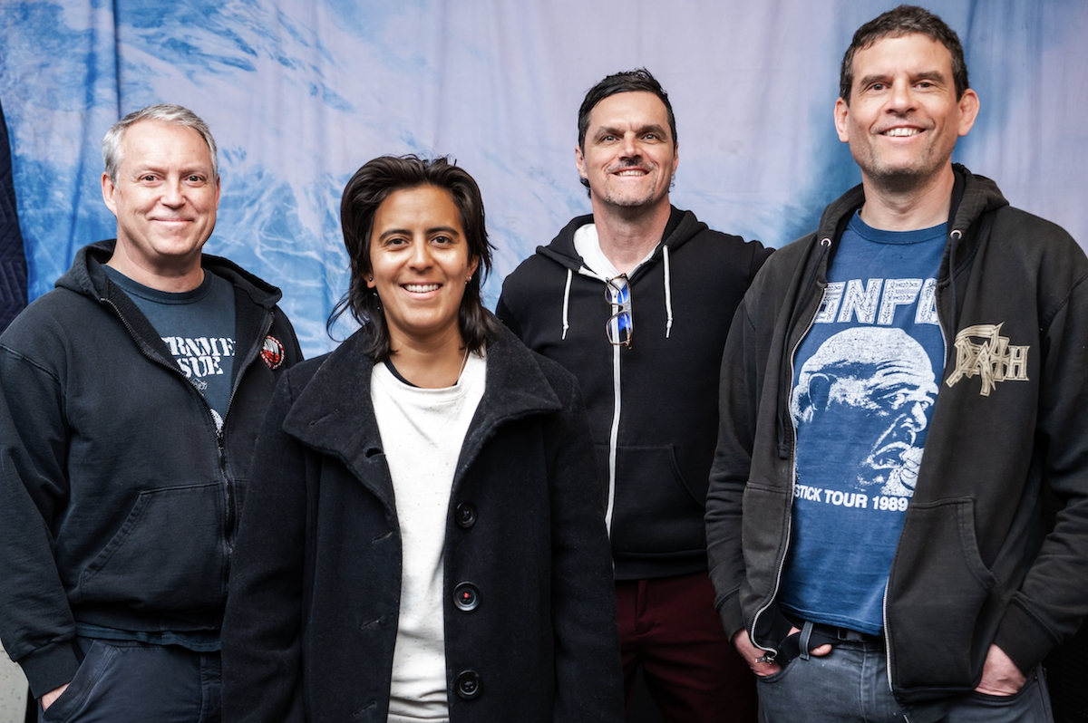 The four members of the punk rock band Propagandhi smile together against a light blue, wave-like backdrop while wearing casual hoodies and t-shirts.