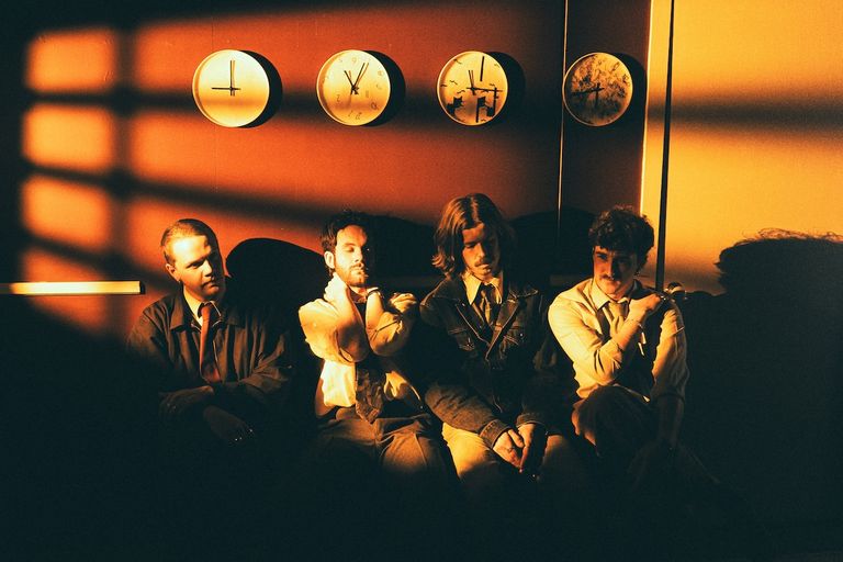 Indie band Purée sitting together under warm orange lighting with four wall clocks above them in a dramatic, cinematic room.