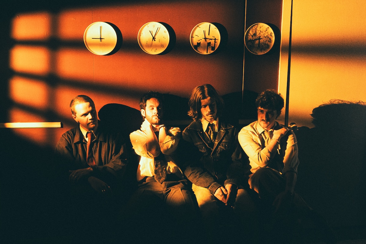 Indie band Purée sitting together under warm orange lighting with four wall clocks above them in a dramatic, cinematic room.