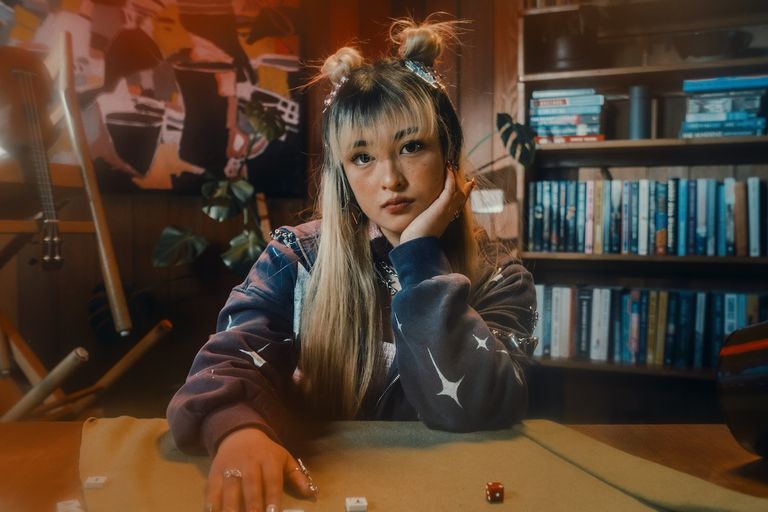 Musician RinRin rests her chin on her hand in a cozy room with bookshelves, sporting space buns hairstyle and a star-pattern jacket.