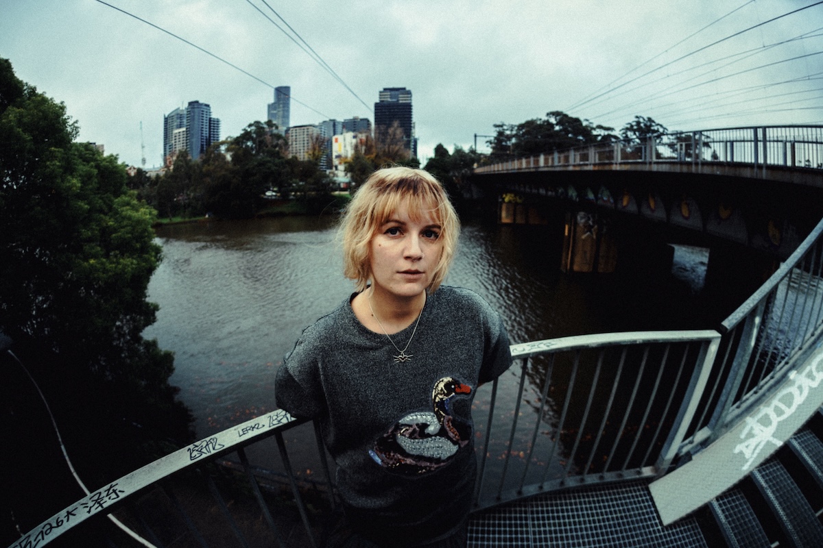 Musician Romanie with short blonde hair and a swan sweater stands on a bridge overlooking a river with a city skyline under a cloudy sky.