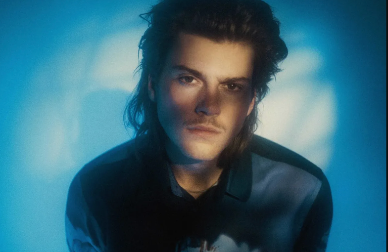 Australian pop singer-songwriter Ruel looking directly forward illuminated by moody blue studio lighting for a promotional artist portrait photo shoot.