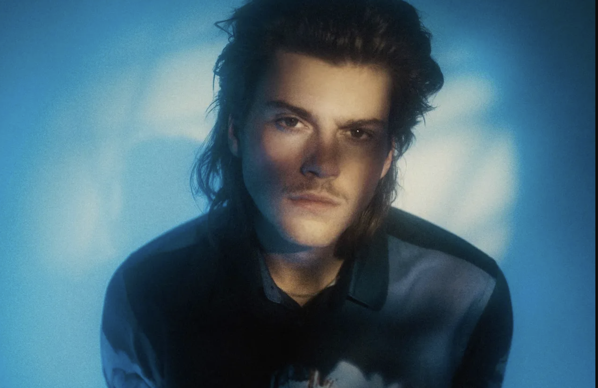 Australian pop singer-songwriter Ruel looking directly forward illuminated by moody blue studio lighting for a promotional artist portrait photo shoot.