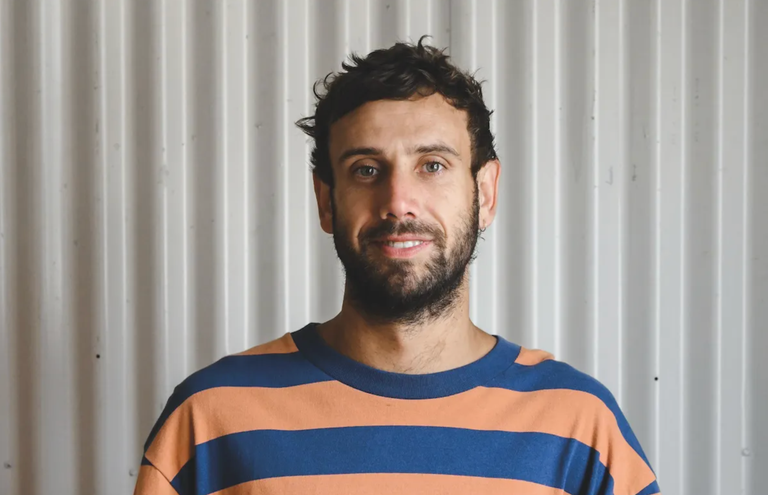 First Nations musical artist Rulla posing in a blue and orange striped shirt against a corrugated metal wall portrait.