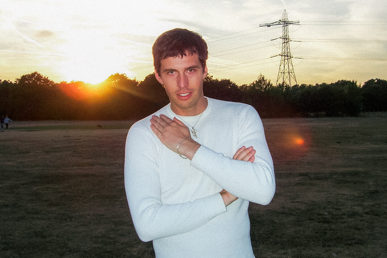 Studio portrait of artist SWIM wearing a white textured long-sleeve shirt outdoors in a park at sunset with an electricity pylon behind