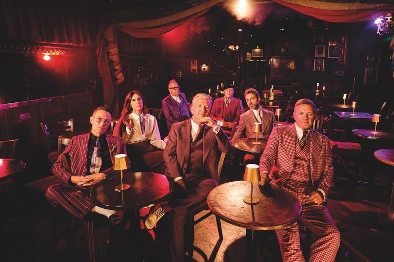 English new wave rock band Squeeze in sharp tailored suits sitting at dimly lit tables in a vintage cabaret club.