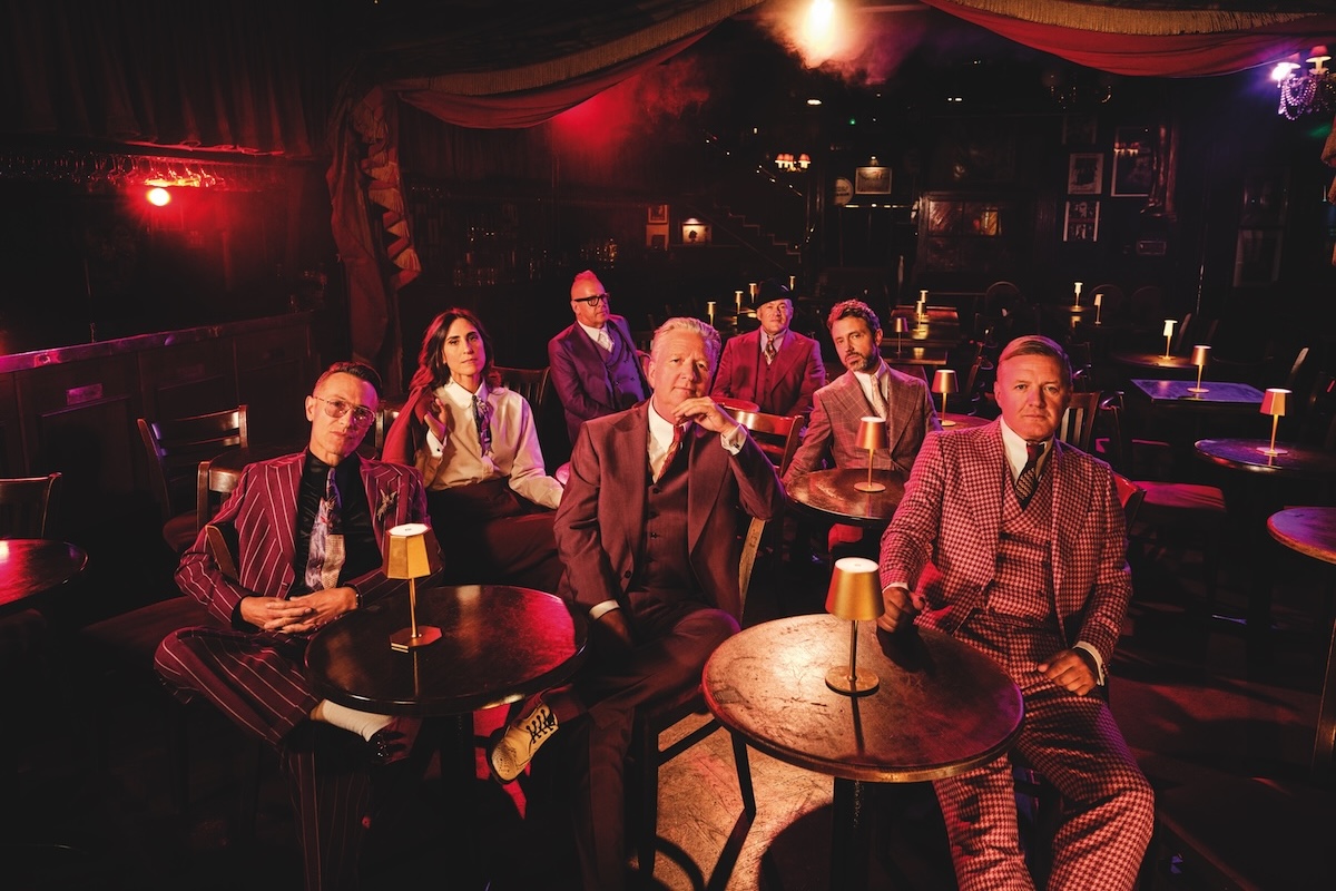 English new wave rock band Squeeze in sharp tailored suits sitting at dimly lit tables in a vintage cabaret club.